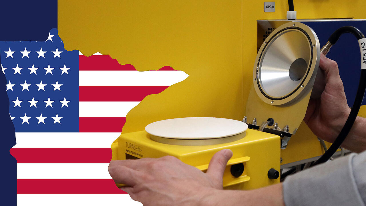 Flag of the U.S. state of Minnesota in the foreground; a hand holding a yellow mask filter tester in the background